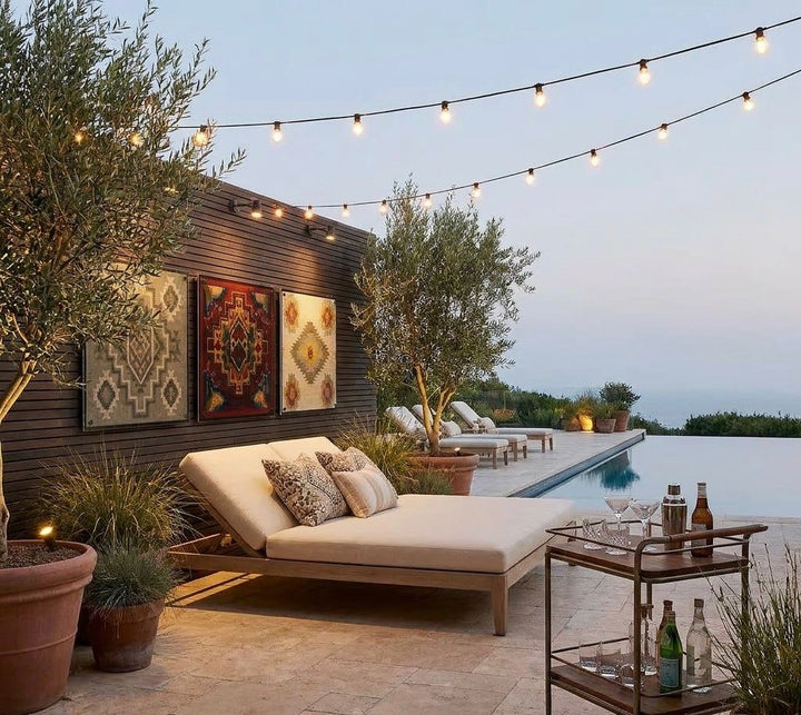 Outdoor patio with lounge chair, decorative pillows, and string lights by a poolside.