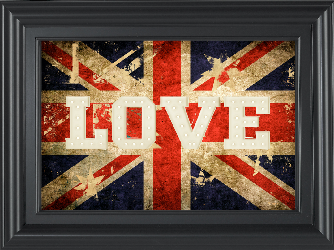 An outdoor setting featuring a large acrylic print with the word 'LOVE' displayed in bold letters against a weathered Union Jack flag backdrop, housed in a black frame. The print is accompanied by potted plants and outdoor furniture on a stone patio.